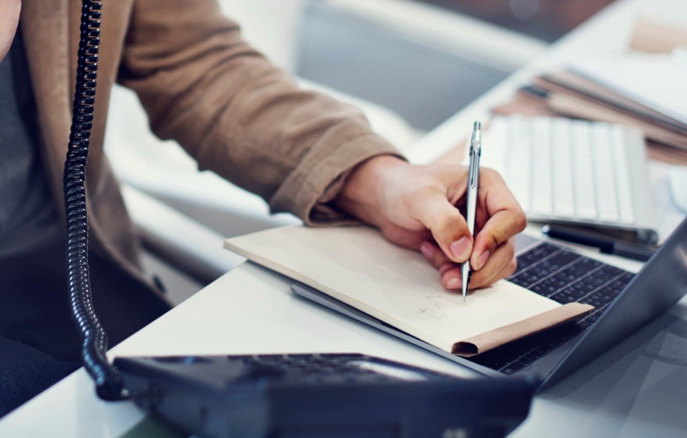 Person taking notes at a desk beside a desk phone and laptop